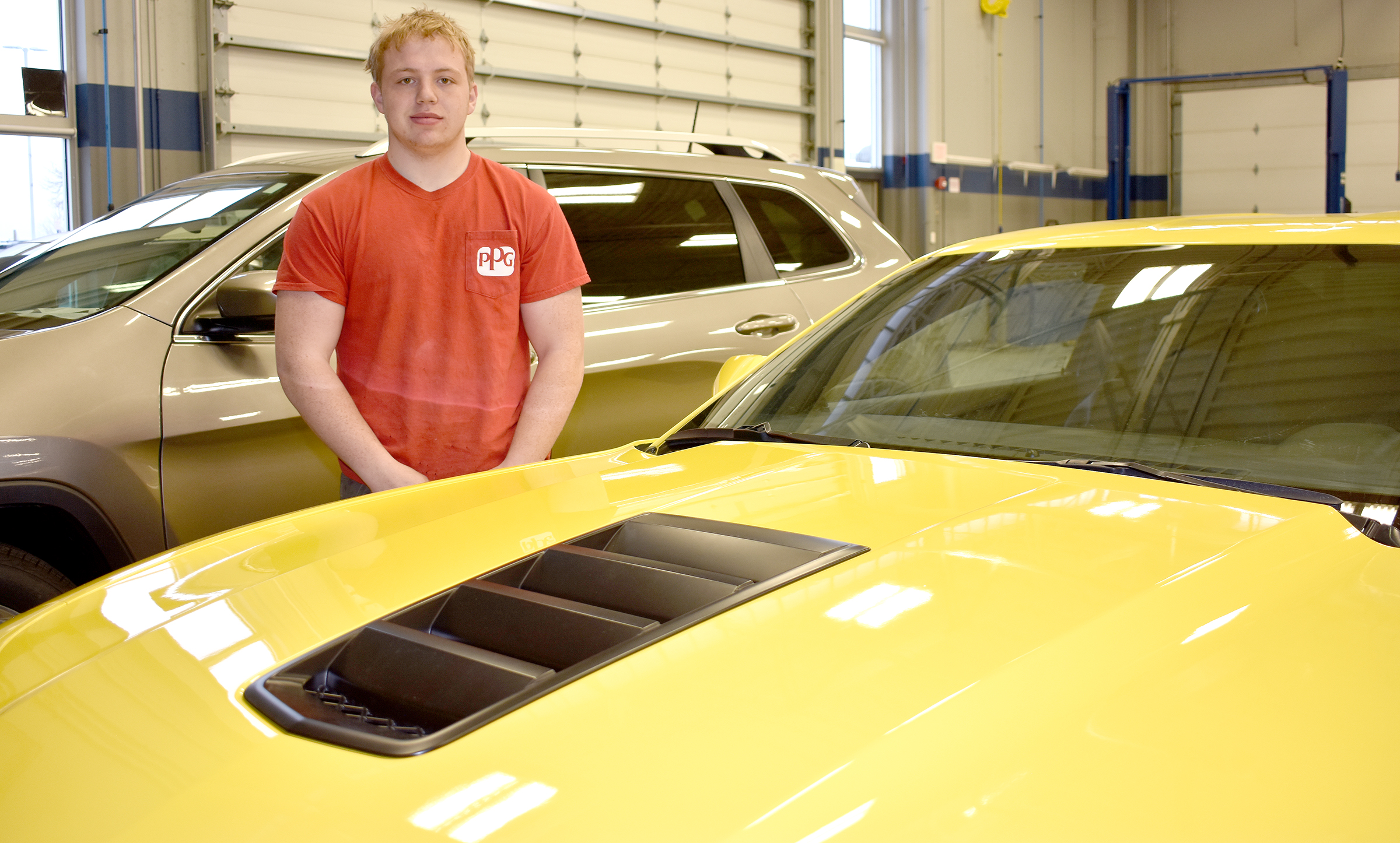 Male student wearing red shirt standing behind yellow Camaro.