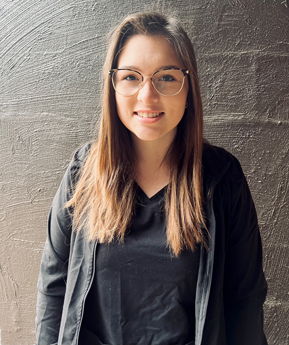 Female with long hair and glasses standing in front of wall.