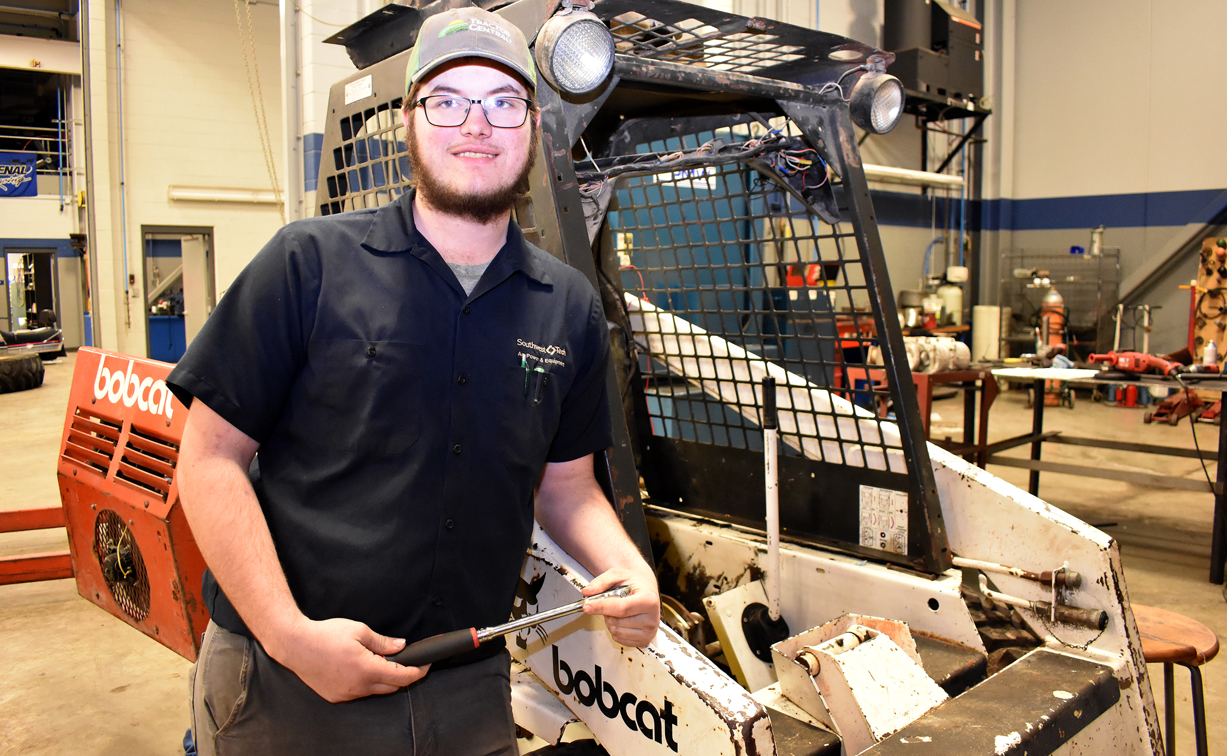 Male student holding wrench standing in front of bobcat vehicle.