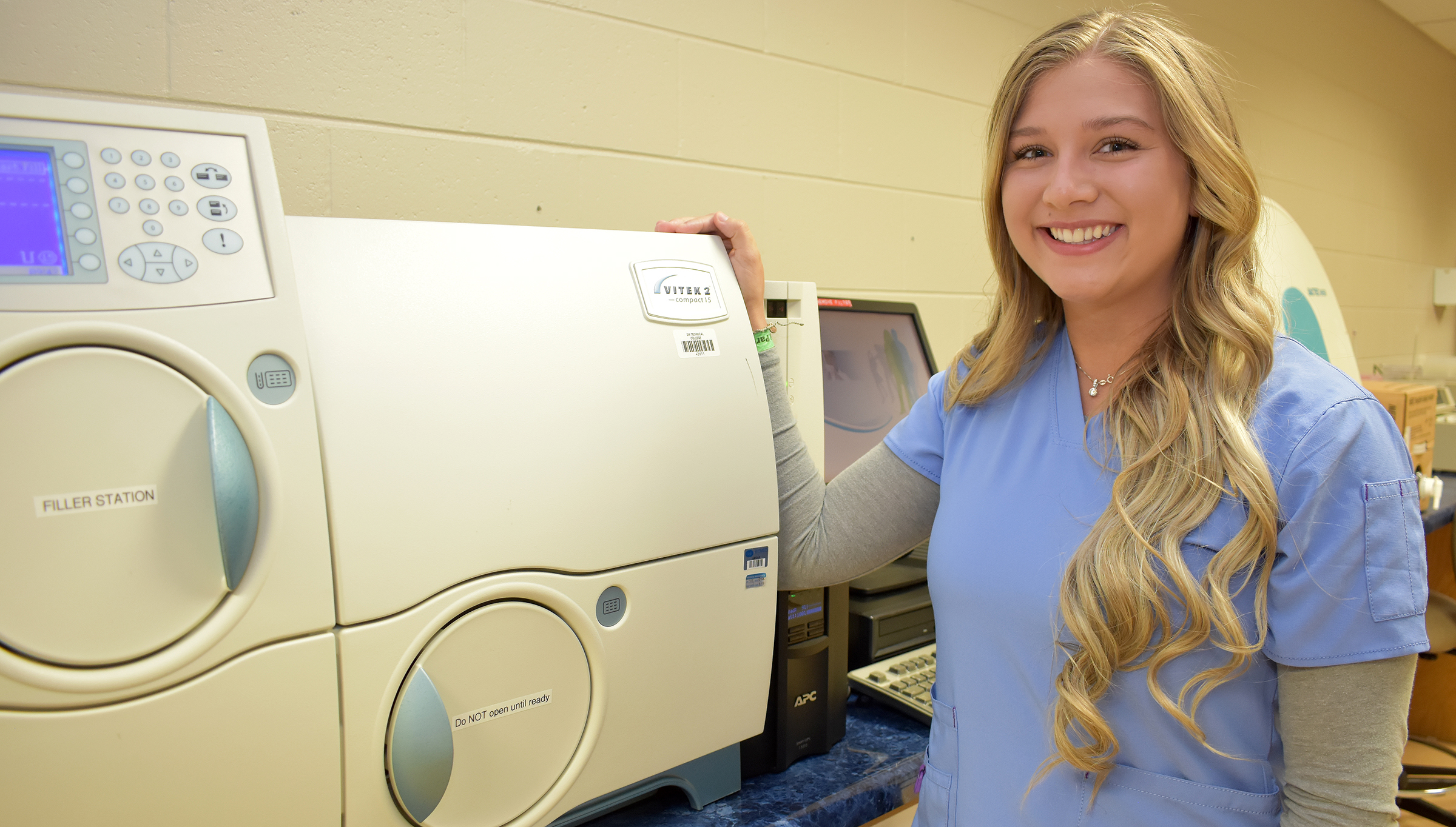 Female student in blue scrubs standing next to medical lab equipment.